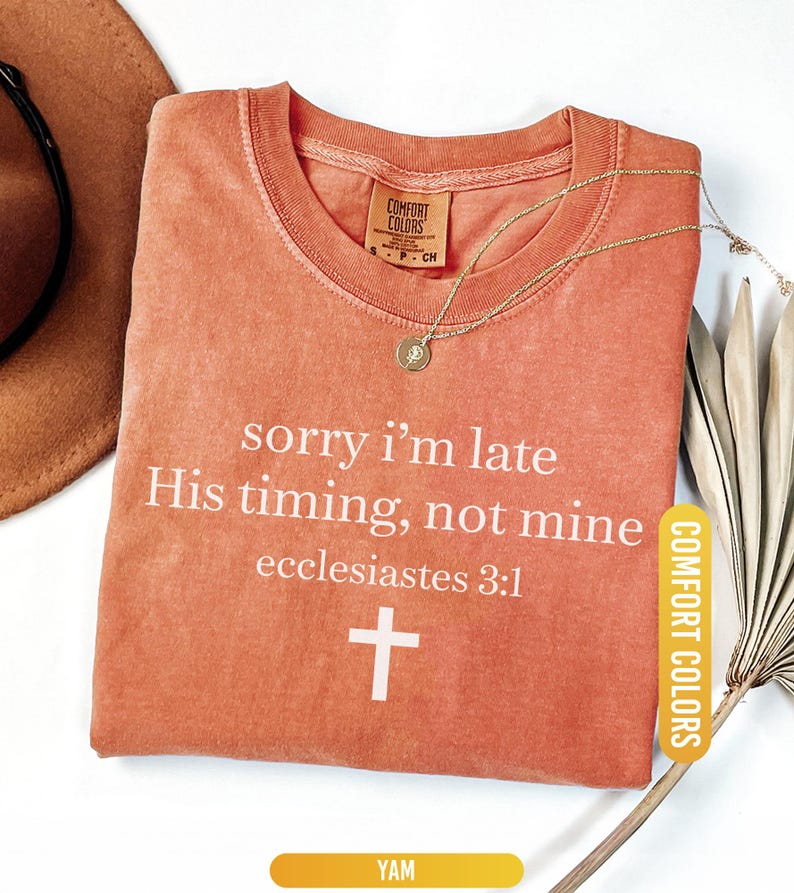 May include: A yam-colored t-shirt with the text "sorry i'm late His timing, not mine ecclesiastes 3:1" and a white cross. A gold necklace and a brown hat are also visible.