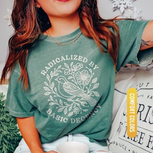 May include: Light green t-shirt with the text "RADICALIZED BY BASIC DECENCY" in a white floral and bird design. The shirt is a comfort color and is light green.