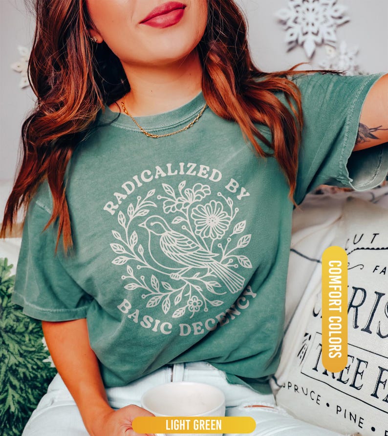 May include: Light green t-shirt with the text "RADICALIZED BY BASIC DECENCY" in a white floral and bird design. The shirt is a comfort color and is light green.