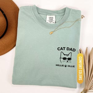 May include: A light green t-shirt with the words "CAT DAD" and a cat wearing sunglasses. The names "MILLIE" and "OLLIE" are below the cat graphic. The shirt is labeled "COMFORT COLORS" and the color is "BAY".