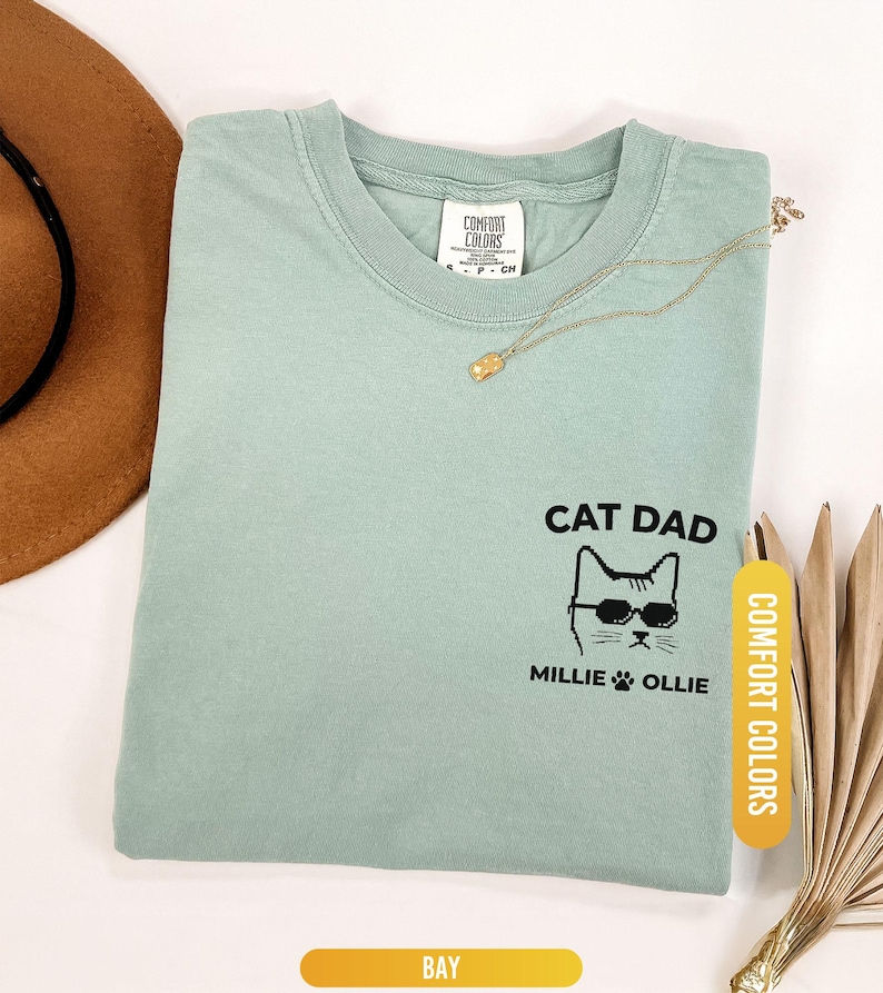 May include: A light green t-shirt with the words "CAT DAD" and a cat wearing sunglasses. The names "MILLIE" and "OLLIE" are below the cat graphic. The shirt is labeled "COMFORT COLORS" and the color is "BAY".