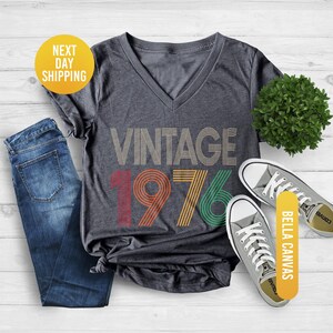 May include: Dark gray heather V-neck t-shirt with the text "VINTAGE 1976" in a retro design. The shirt is paired with blue jeans and gray canvas sneakers. The shirt is made by Bella Canvas.