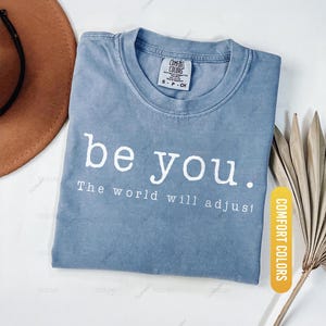 May include: A blue jean-colored t-shirt with the words "be you. The world will adjust" printed in white. The shirt is folded and has a tag that says "Comfort Colors". A brown hat and dried leaves are in the background.