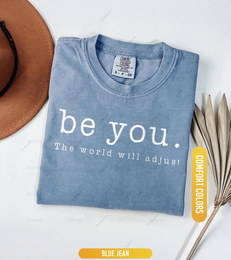 May include: A blue jean-colored t-shirt with the words "be you. The world will adjust" printed in white. The shirt is folded and has a tag that says "Comfort Colors". A brown hat and dried leaves are in the background.