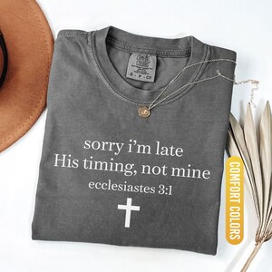 May include: A gray Comfort Colors t-shirt with the text "sorry i'm late His timing, not mine ecclesiastes 3:1" and a white cross. A gold necklace and a brown hat are visible. The shirt is labeled "Pepper".