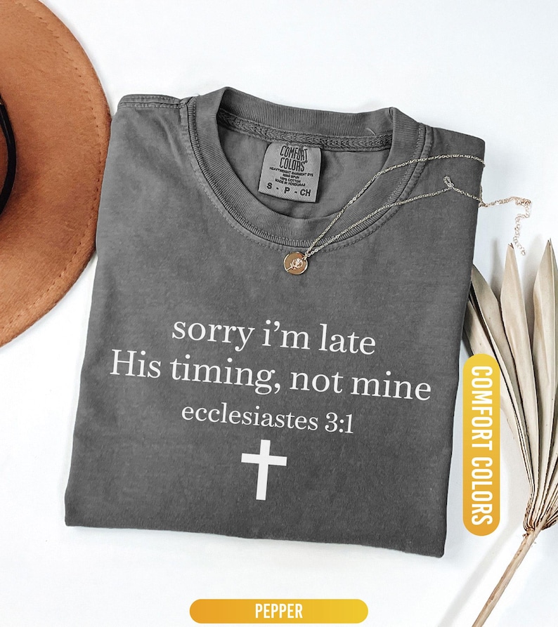 May include: A gray Comfort Colors t-shirt with the text "sorry i'm late His timing, not mine ecclesiastes 3:1" and a white cross. A gold necklace and a brown hat are visible. The shirt is labeled "Pepper".