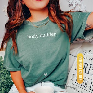 Pu&ograve; includere: T-shirt verde chiaro con la scritta "body builder" in bianco. La maglietta ha un piccolo cuore bianco. La maglietta &egrave; un colore confortevole ed &egrave; verde chiaro.