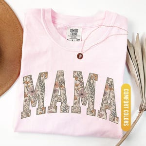 Op de afbeelding: Een lichtroze t-shirt met het woord "MAMA" in grote letters met bloemenpatroon. Het t-shirt heeft een ronde hals en korte mouwen. Een gouden ketting met een kleine hanger ligt op het shirt. Het shirt is gelabeld met "Blossom" en "Comfort Colors".