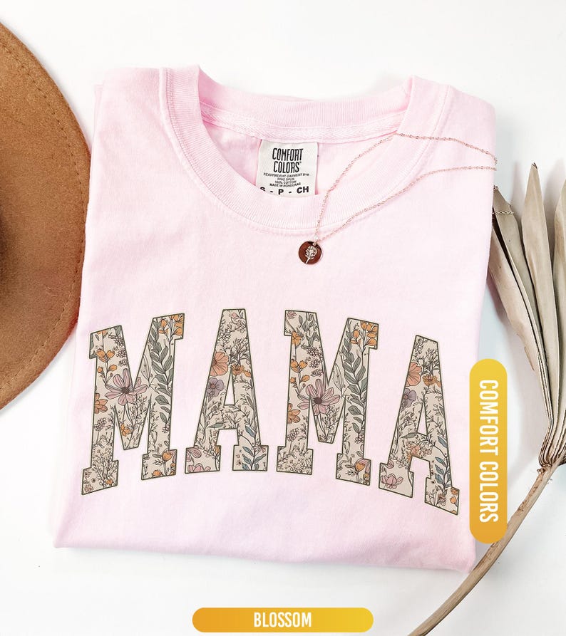 Op de afbeelding: Een lichtroze t-shirt met het woord "MAMA" in grote letters met bloemenpatroon. Het t-shirt heeft een ronde hals en korte mouwen. Een gouden ketting met een kleine hanger ligt op het shirt. Het shirt is gelabeld met "Blossom" en "Comfort Colors".