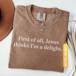 May include: Espresso-colored t-shirt with the text "First of all, Jesus thinks I'm a delight." The shirt has a crew neck and a Comfort Colors tag. A silver necklace with a pendant is draped on the shirt.