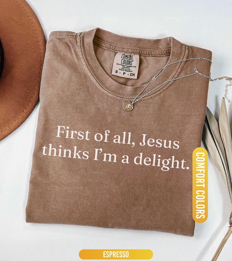May include: Espresso-colored t-shirt with the text "First of all, Jesus thinks I'm a delight." The shirt has a crew neck and a Comfort Colors tag. A silver necklace with a pendant is draped on the shirt.