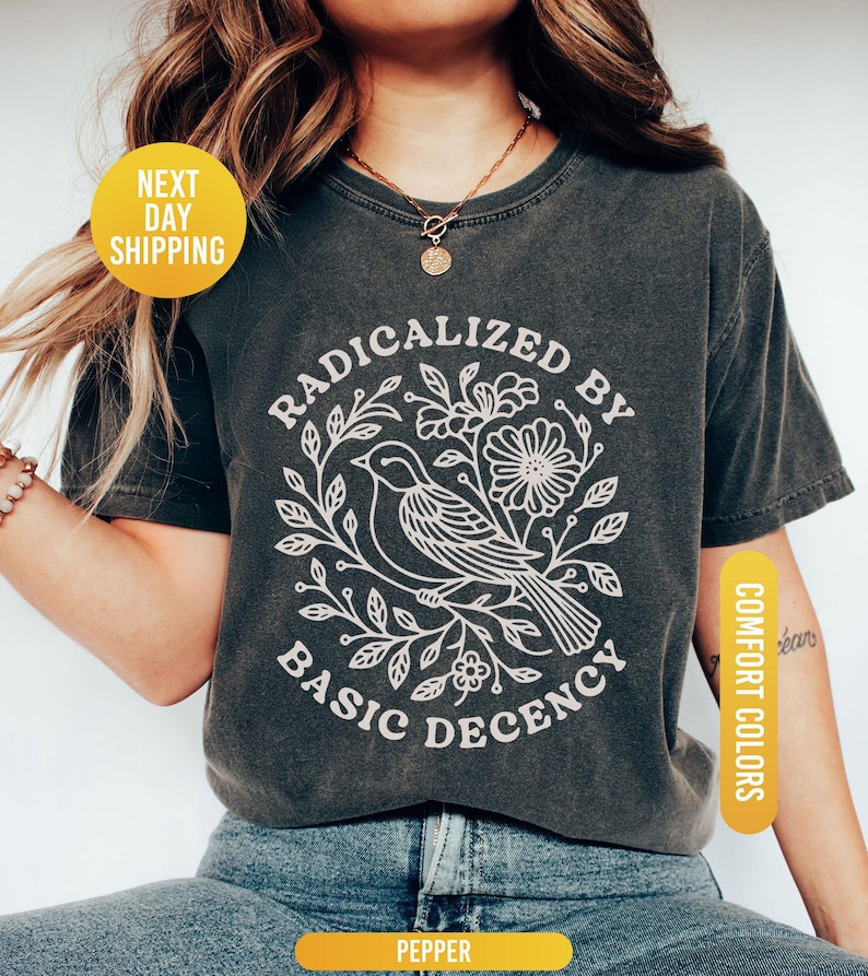 May include: Dark grey t-shirt with a white graphic of a bird surrounded by flowers and leaves, with the text "RADICALIZED BY BASIC DECENCY". The shirt has a round neck and short sleeves. The shirt is labeled "Pepper" and "Comfort Colors".
