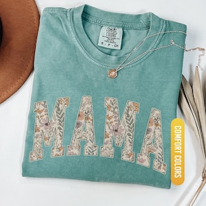 Op de afbeelding: Lichtgroen t-shirt met het woord "MAMA" in een bloemenpatroon. Het shirt heeft een ronde hals en korte mouwen. Een gouden ketting met een hanger ligt op het shirt. Het shirt is voorzien van het label "Comfort Colors".