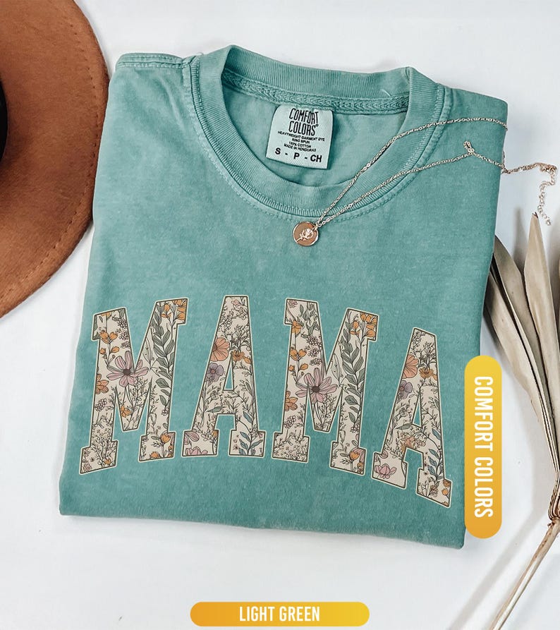 Op de afbeelding: Lichtgroen t-shirt met het woord "MAMA" in een bloemenpatroon. Het shirt heeft een ronde hals en korte mouwen. Een gouden ketting met een hanger ligt op het shirt. Het shirt is voorzien van het label "Comfort Colors".