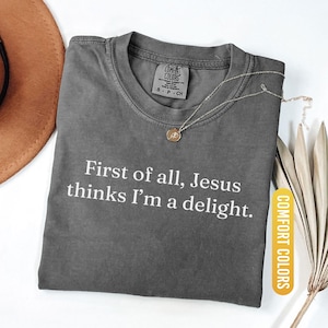 May include: Pepper-colored t-shirt with the text "First of all, Jesus thinks I'm a delight." A gold necklace with a pendant rests on the shirt. The shirt is labeled "Comfort Colors."