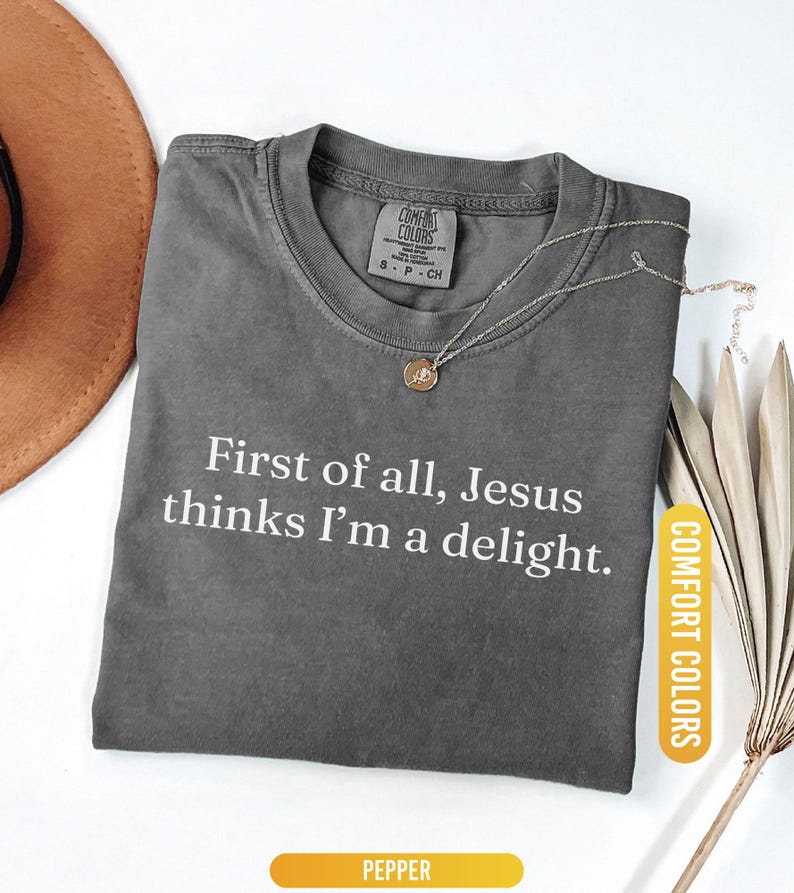 May include: Pepper-colored t-shirt with the text "First of all, Jesus thinks I'm a delight." A gold necklace with a pendant rests on the shirt. The shirt is labeled "Comfort Colors."