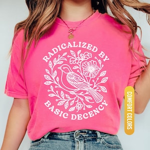 May include: A bright pink t-shirt with the text "RADICALIZED BY BASIC DECENCY" in a white circular design featuring a bird and floral elements. The shirt is a comfort color and is paired with blue jeans.