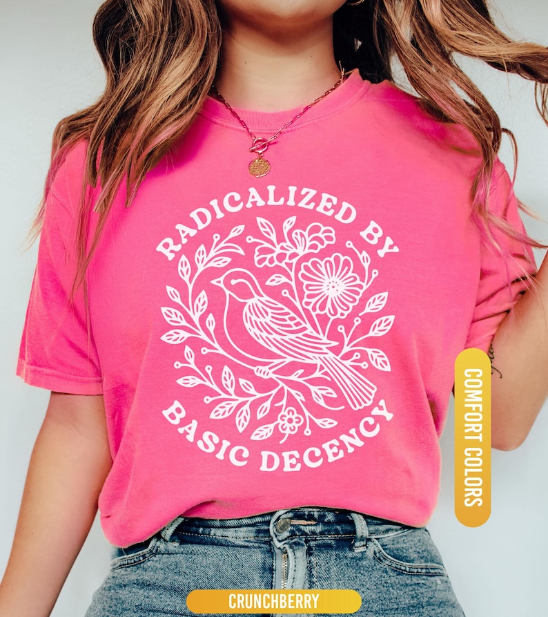 May include: A bright pink t-shirt with the text "RADICALIZED BY BASIC DECENCY" in a white circular design featuring a bird and floral elements. The shirt is a comfort color and is paired with blue jeans.