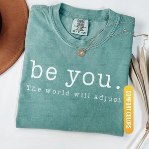 May include: Light green Comfort Colors t-shirt with the text "be you. The world will adjust" in white. A gold necklace with a pendant rests on the shirt. A brown hat is partially visible.