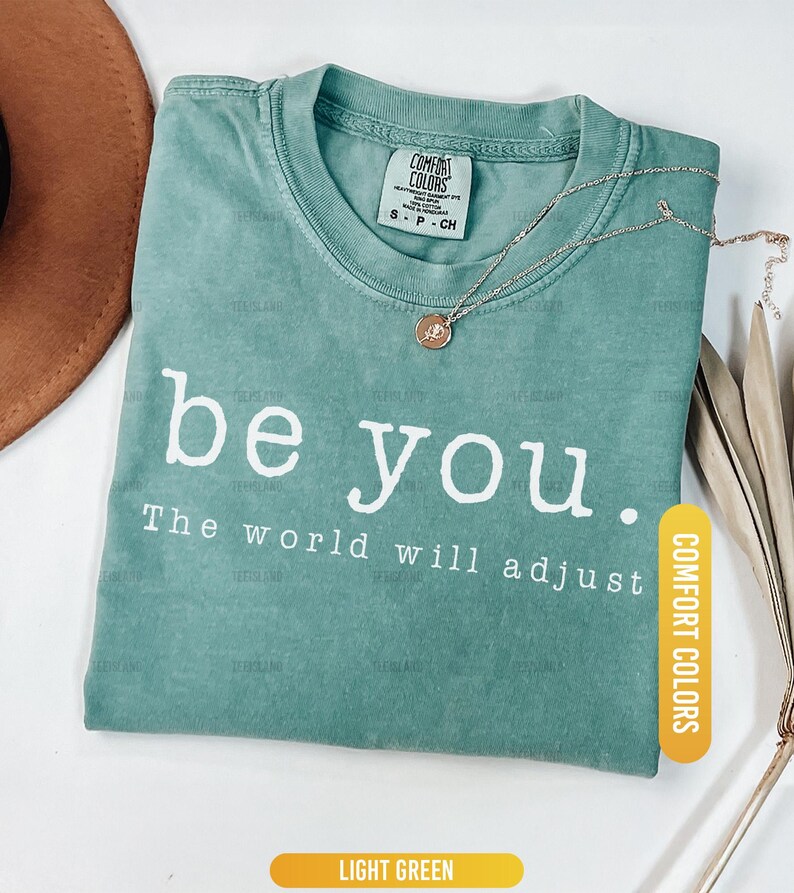 May include: Light green Comfort Colors t-shirt with the text "be you. The world will adjust" in white. A gold necklace with a pendant rests on the shirt. A brown hat is partially visible.