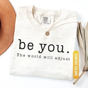 May include: Ivory-colored t-shirt with the text "be you. The world will adjust" in black. A gold necklace with a pendant rests on the shirt. A brown hat and dried leaves are in the background.
