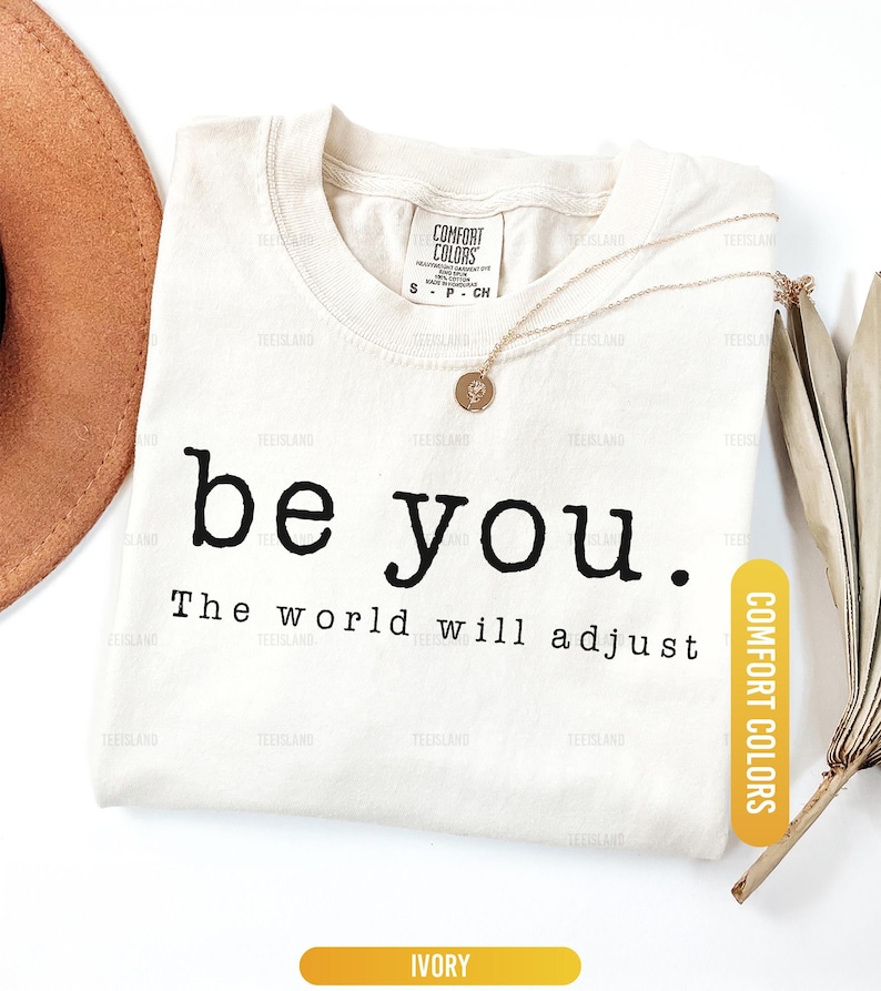 May include: Ivory-colored t-shirt with the text "be you. The world will adjust" in black. A gold necklace with a pendant rests on the shirt. A brown hat and dried leaves are in the background.