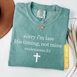 May include: Light green t-shirt with the text "sorry i'm late His timing, not mine ecclesiastes 3:1" and a white cross. A gold necklace with a pendant is on the shirt. The shirt is labeled "Comfort Colors".