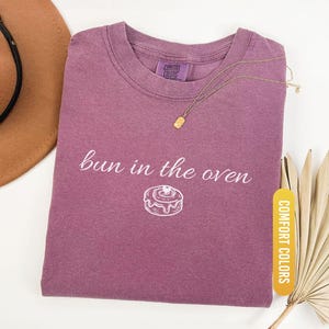 May include: Berry-colored t-shirt with the phrase "bun in the oven" and a graphic of a donut. A gold necklace with a rectangular pendant rests on the shirt. The shirt is labeled "Comfort Colors".