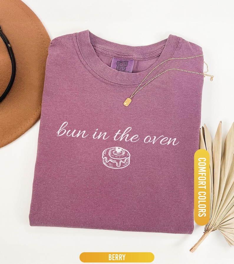 May include: Berry-colored t-shirt with the phrase "bun in the oven" and a graphic of a donut. A gold necklace with a rectangular pendant rests on the shirt. The shirt is labeled "Comfort Colors".
