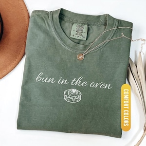 May include: Moss green t-shirt with the phrase "bun in the oven" and a cinnamon roll graphic. The shirt has a crew neck and short sleeves. A gold necklace with a small pendant rests on the shirt. The shirt is labeled "Comfort Colors".