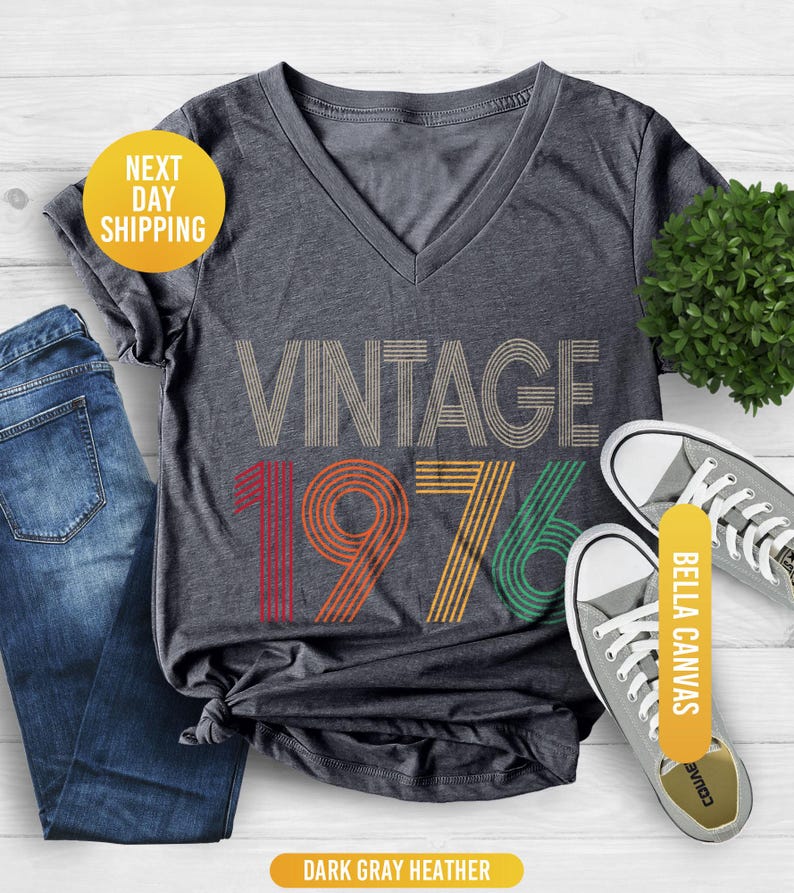 May include: Dark gray heather V-neck t-shirt with the text "VINTAGE 1976" in a retro design. The shirt is paired with blue jeans and gray canvas sneakers. The shirt has a knot at the bottom.