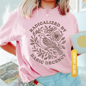 May include: A light pink t-shirt with the words "RADICALIZED BY BASIC DECENCY" surrounding a bird and floral design in brown. The shirt is labeled "Blossom" and "Comfort Colors".