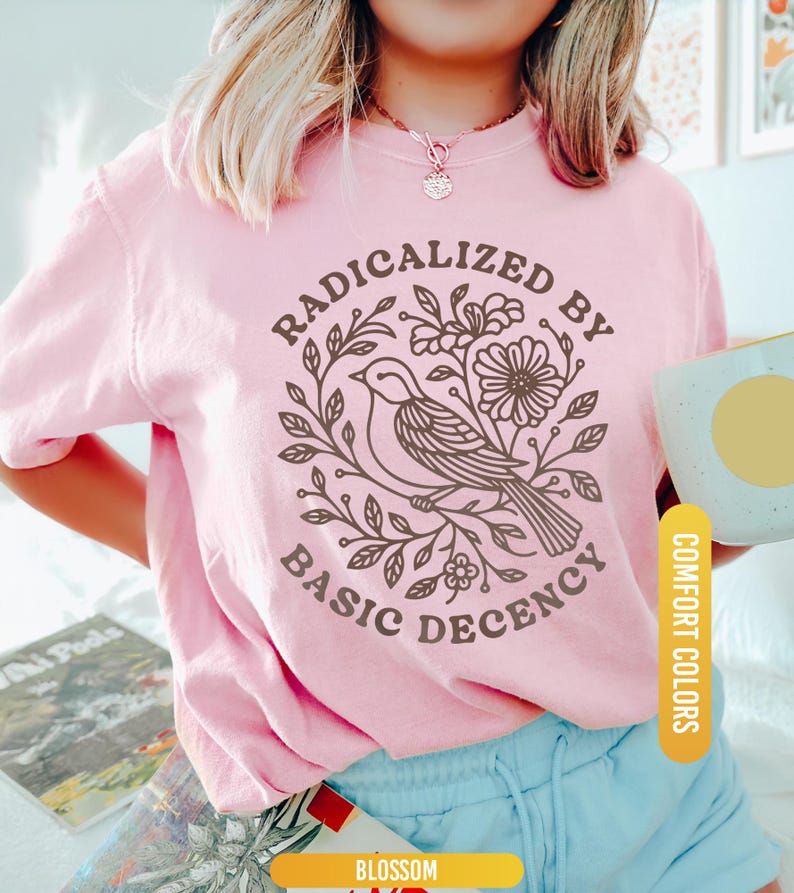 May include: A light pink t-shirt with the words "RADICALIZED BY BASIC DECENCY" surrounding a bird and floral design in brown. The shirt is labeled "Blossom" and "Comfort Colors".