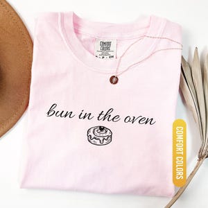 May include: A light pink t-shirt with the phrase "bun in the oven" and a graphic of a cinnamon roll. The shirt is a Comfort Colors brand in the color Blossom. A gold necklace with a pendant is also visible.