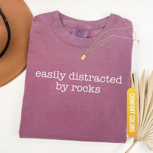 May include: Berry-colored t-shirt with the phrase "easily distracted by rocks" in white lettering. A gold necklace with a rectangular pendant rests on the shirt. The shirt is labeled "Comfort Colors".