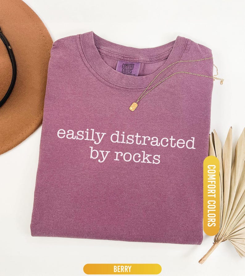 May include: Berry-colored t-shirt with the phrase "easily distracted by rocks" in white lettering. A gold necklace with a rectangular pendant rests on the shirt. The shirt is labeled "Comfort Colors".