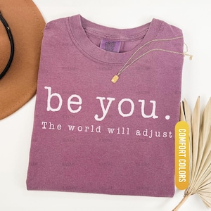May include: Berry-colored t-shirt with the words "be you." and "The world will adjust" printed in white. A gold necklace with a rectangular pendant rests on the shirt. The shirt is labeled "Comfort Colors".