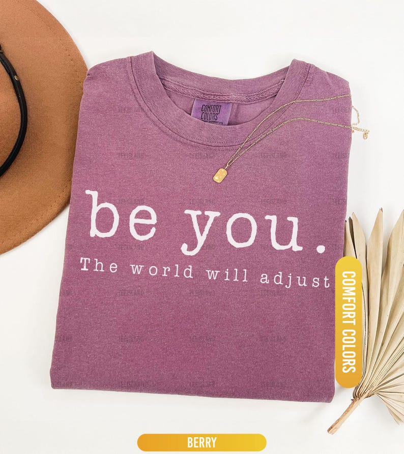 May include: Berry-colored t-shirt with the words "be you." and "The world will adjust" printed in white. A gold necklace with a rectangular pendant rests on the shirt. The shirt is labeled "Comfort Colors".