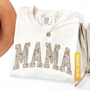 Op de afbeelding: Ivoor kleurig T-shirt met het woord "MAMA" in grote letters met een bloemenpatroon. Het shirt heeft een ronde hals en een klein label bij de halslijn. Een gouden ketting en een bruine hoed zijn ook zichtbaar. Het shirt is gelabeld "Comfort Colors".