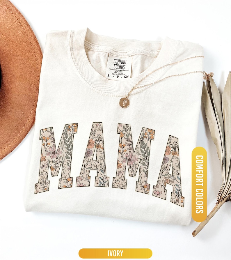 Op de afbeelding: Ivoor kleurig T-shirt met het woord "MAMA" in grote letters met een bloemenpatroon. Het shirt heeft een ronde hals en een klein label bij de halslijn. Een gouden ketting en een bruine hoed zijn ook zichtbaar. Het shirt is gelabeld "Comfort Colors".