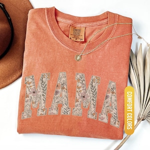 Op de afbeelding: Een koraalkleurig t-shirt met het woord "MAMA" in grote, bloemmotief letters. Het shirt heeft een ronde hals en korte mouwen. Een gouden ketting met een hanger is over het shirt gedrapeerd. Het shirt is gelabeld "Comfort Colors".