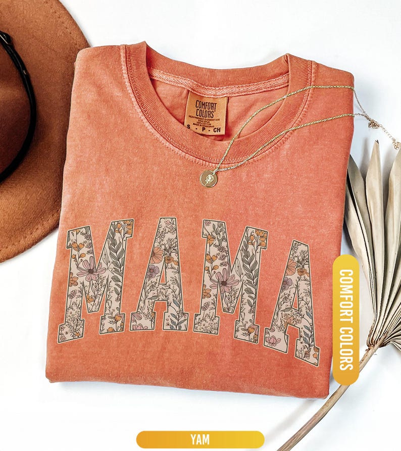 Op de afbeelding: Een koraalkleurig t-shirt met het woord "MAMA" in grote, bloemmotief letters. Het shirt heeft een ronde hals en korte mouwen. Een gouden ketting met een hanger is over het shirt gedrapeerd. Het shirt is gelabeld "Comfort Colors".