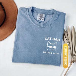 May include: Blue jean colored t-shirt with the words "CAT DAD" and a cat wearing sunglasses. The names "Millie & Ollie" and a paw print are below the cat graphic. The shirt is labeled "Comfort Colors".