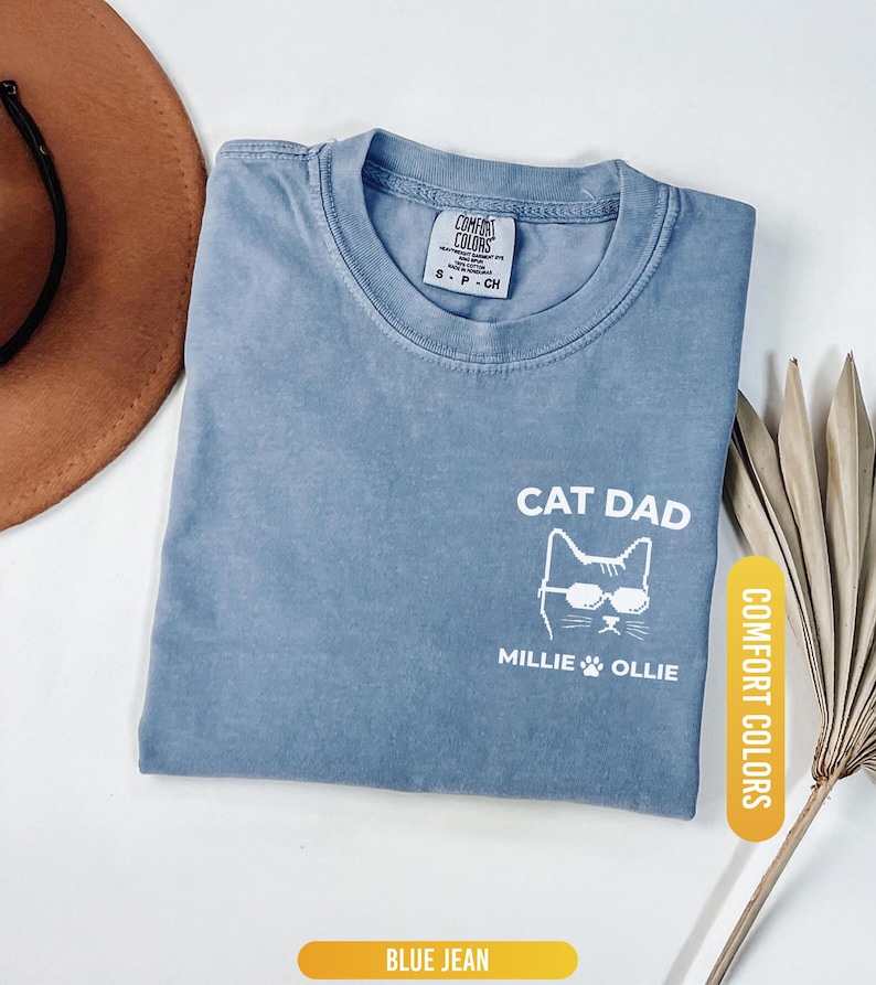 May include: Blue jean colored t-shirt with the words "CAT DAD" and a cat wearing sunglasses. The names "Millie & Ollie" and a paw print are below the cat graphic. The shirt is labeled "Comfort Colors".