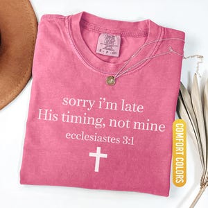 May include: A pink Comfort Colors t-shirt with the text "sorry i'm late His timing, not mine ecclesiastes 3:1" and a white cross. A gold-colored necklace with a pendant is on the shirt. The shirt is labeled "Crunchberry".