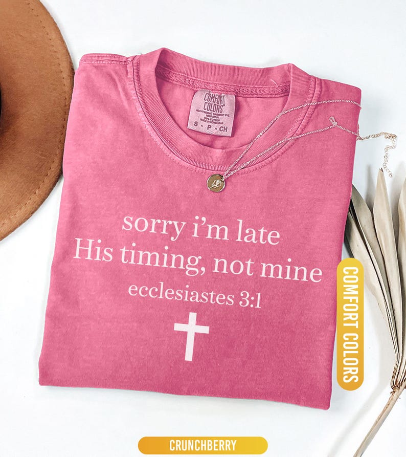 May include: A pink Comfort Colors t-shirt with the text "sorry i'm late His timing, not mine ecclesiastes 3:1" and a white cross. A gold-colored necklace with a pendant is on the shirt. The shirt is labeled "Crunchberry".