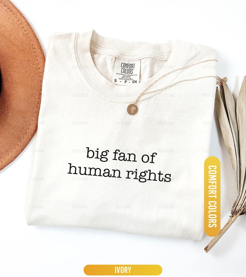 May include: Ivory-colored t-shirt with the text "big fan of human rights" in black. A gold necklace with a pendant rests on the shirt. The shirt has a Comfort Colors tag and an ivory color label.