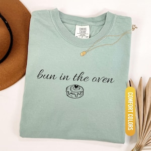 May include: A light green t-shirt with the phrase "bun in the oven" and a graphic of a cinnamon roll. The shirt has a crew neck and a tag that says "Comfort Colors". A gold necklace and a brown hat are also in the image.