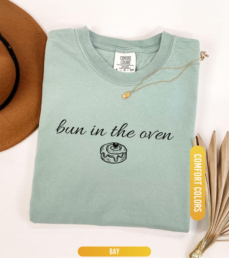 May include: A light green t-shirt with the phrase "bun in the oven" and a graphic of a cinnamon roll. The shirt has a crew neck and a tag that says "Comfort Colors". A gold necklace and a brown hat are also in the image.