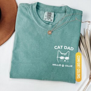 May include: Light green t-shirt with the text "CAT DAD" and a cat graphic wearing sunglasses. The shirt also features the names "Millie" and "Ollie" below the cat graphic. A gold necklace and a brown hat are also visible.